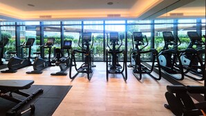 A gym with multiple cardio machines, a weight bench, and large windows offering a view of greenery and buildings.