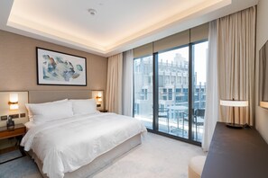 A modern hotel room with a large bed, a city view through floor-to-ceiling windows, a ceiling light, and a framed abstract painting.