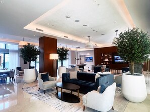 A modern hotel lobby with a seating area, a dining area, and a kitchen in the background.