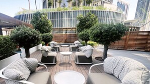 A rooftop lounge with modern outdoor furniture, including sofas and a round table, surrounded by greenery and overlooking a cityscape with high-rise buildings.