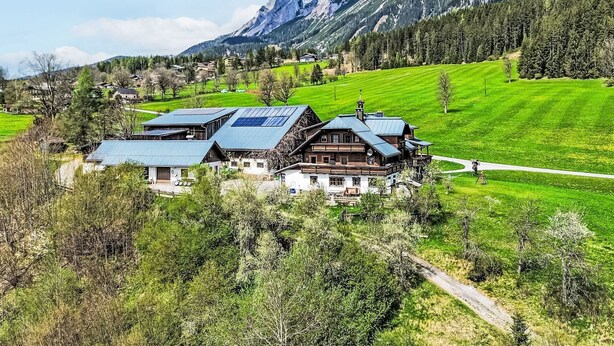 A traditional chalet with a sloped roof, surrounded by lush green fields and mountains in the background.