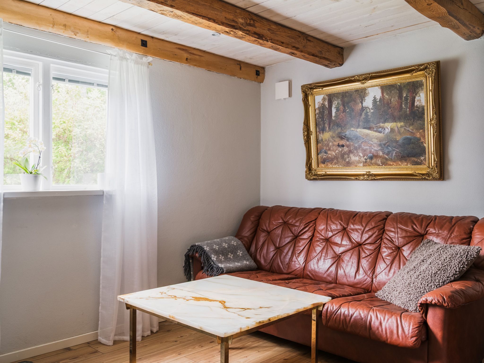 A room with a brown leather sofa, a white marble table, a painting on the wall, and a window with a curtain.