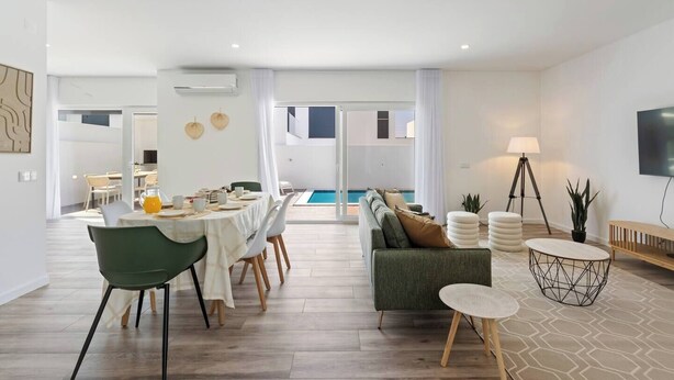 A modern living room with a dining area, a sofa, a TV, and a pool view.