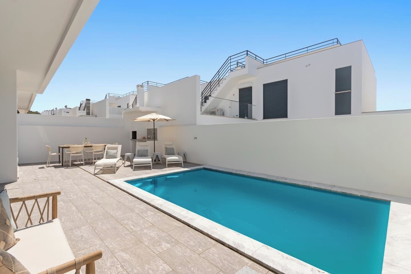 A modern pool area with a rectangular pool, white walls, and a terrace with lounge chairs and a table.