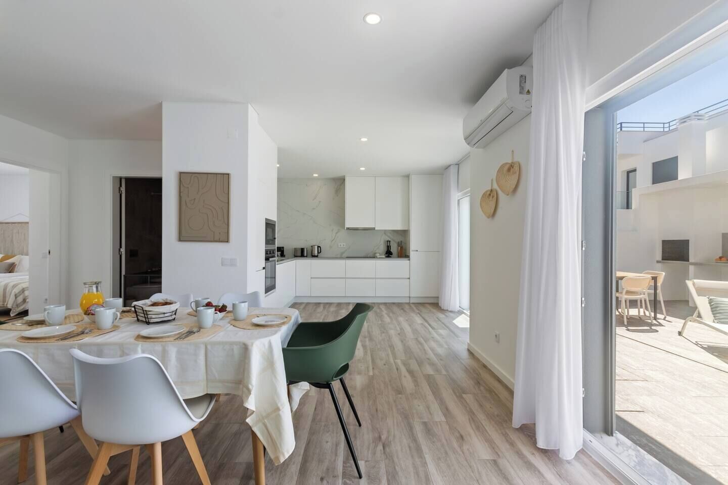 Une cuisine moderne avec des meubles blancs, un coin repas avec une table dressée pour quatre personnes, et une grande porte-fenêtre coulissante donnant sur un espace extérieur aménagé pour se détendre.