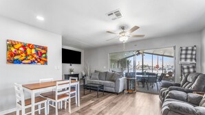 A modern living room with a dining area, grey sofas, a ceiling fan, and a large window offering a view of the outdoors. - Dockside Delight! Waterfront Home + Boat Slip for Your Next Getaway (New Port Richey)