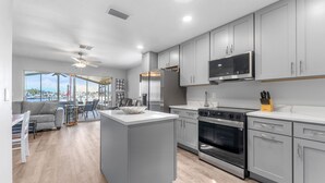 A modern kitchen with grey cabinets, a stainless steel refrigerator, a microwave, and an oven. There is a dining area with a table and chairs, and a view of the marina through large windows. - Dockside Delight! Waterfront Home + Boat Slip for Your Next Getaway (New Port Richey)