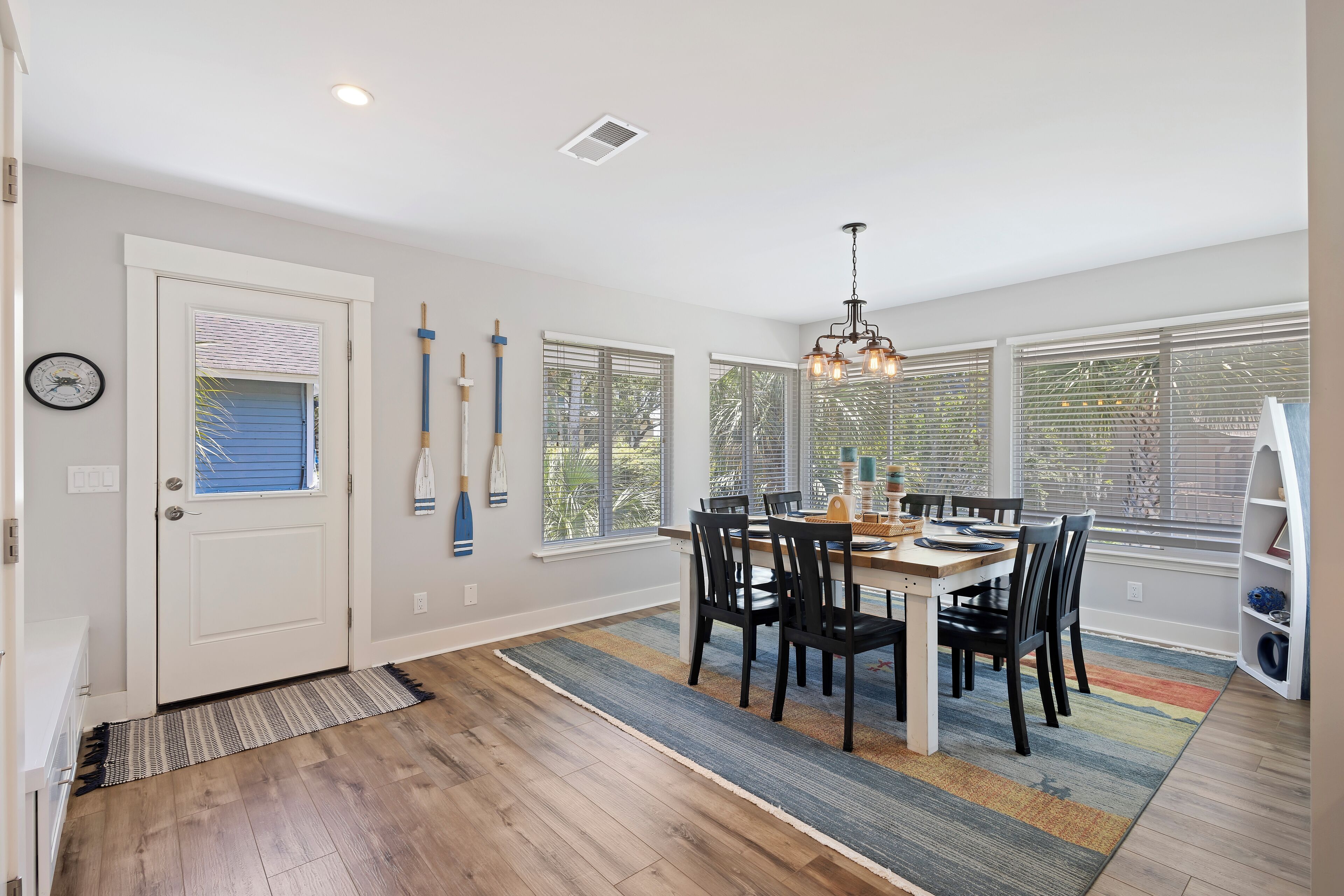 A dining area with a wooden table and chairs, a chandelier, and large windows with blinds.