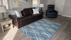 Een woonkamer met een bruine leren bank, een blauw tapijt met patroon, een zwarte leren fauteuil, een kleine tafel met een lamp en een schilderij van surfplanken aan de muur.