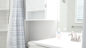 Een badkamer met een wit toilet, een douche met een blauw-wit gestreept douchegordijn, een witte wastafel met een spiegel erboven en witte kastjes.