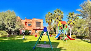 A playground with swings and slides, surrounded by a well-maintained lawn and palm trees.