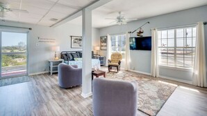 A spacious living room with a large window, a ceiling fan, and a flat-screen TV mounted on the wall. - Deck + Gulf of America View: `Sunrise` on Perdido Key! (Pensacola)