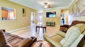 A spacious living room with a brown leather sofa, a green armchair, and a coffee table. A flat-screen TV is mounted on the wall, and there is a ceiling fan above.