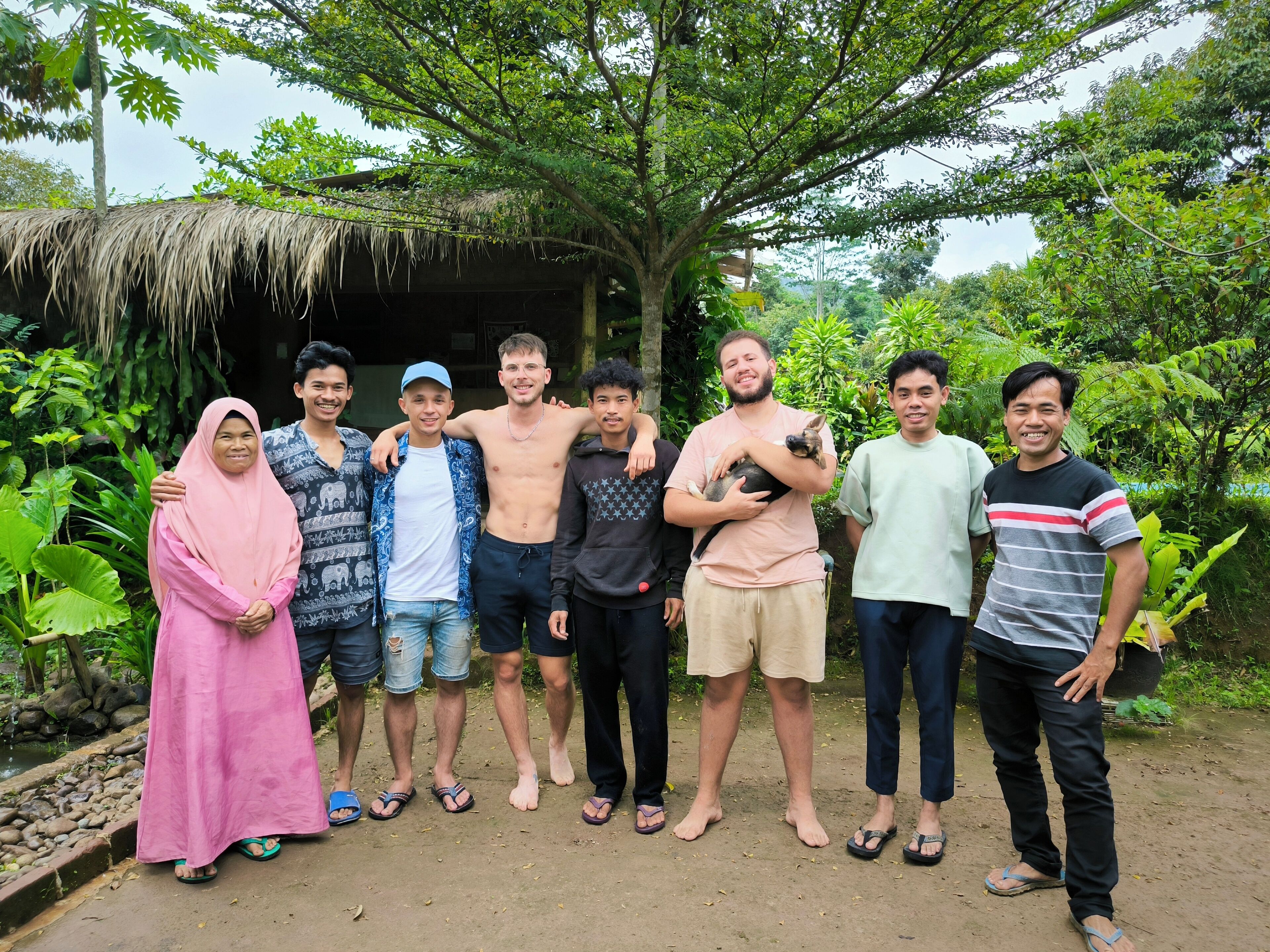 A group of people standing outdoors with a thatched-roof structure and lush greenery in the background.