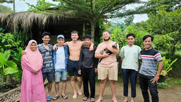 A group of people standing outdoors with a thatched-roof structure and lush greenery in the background.
