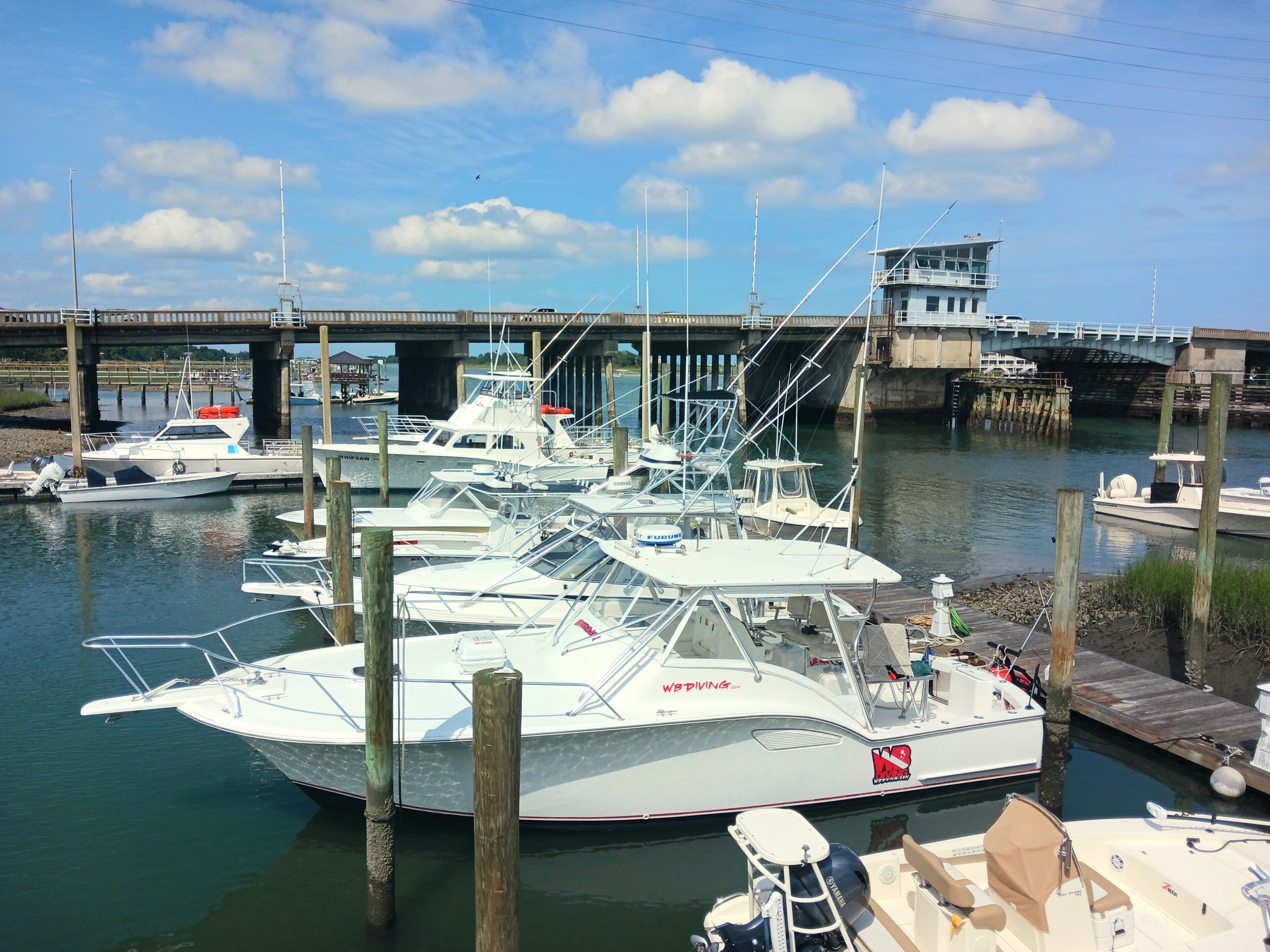 A marina with several boats docked, a bridge in the background, and a wooden dock with a boat on it.