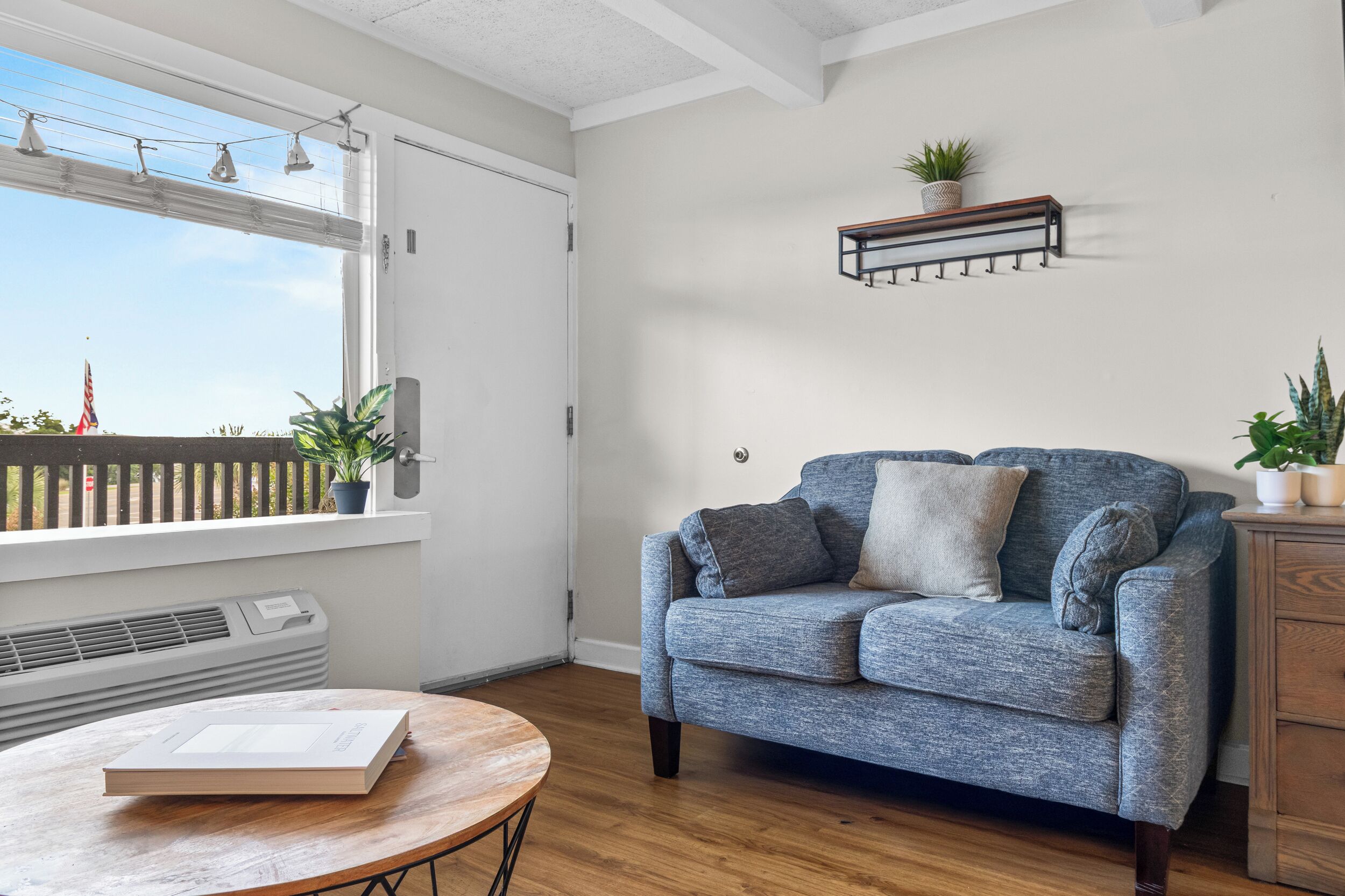 Un salon avec un canapé bleu, une table basse en bois, une fenêtre donnant sur l’extérieur et une étagère à plantes fixée au mur.