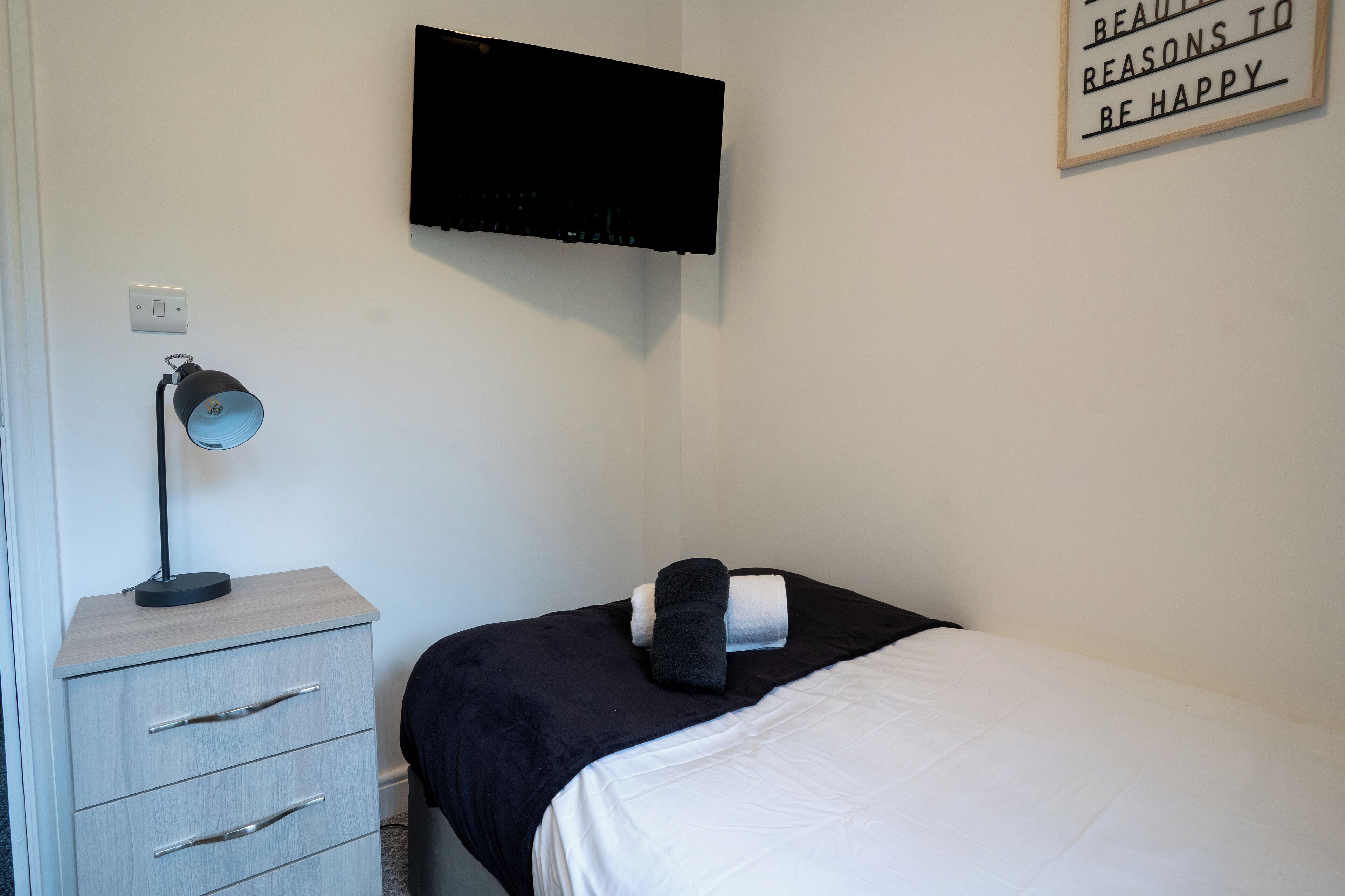 A bedroom with a bed, a bedside table with a lamp, a TV mounted on the wall, and a motivational quote on the wall.