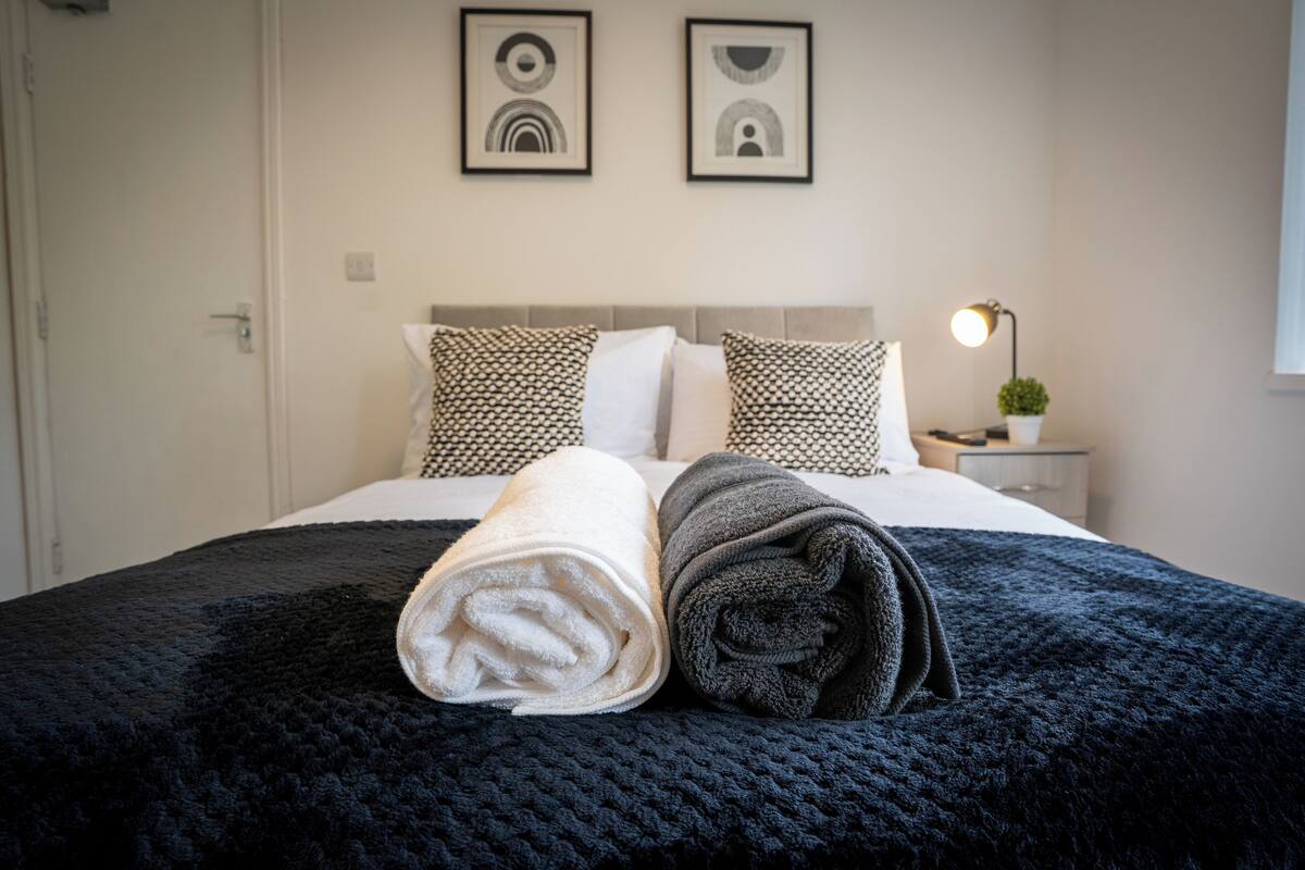 A neatly made bed with a dark blue blanket and two rolled towels, a bedside lamp, and two framed artworks on the wall.