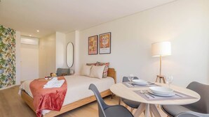 A hotel room with a bed, a dining table with chairs, a lamp, and two framed pictures on the wall.