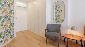 A room with a patterned curtain, a chair, a table with a lamp, and a wall-mounted mirror.