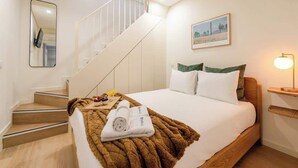 A hotel room with a bed, a wooden headboard, a nightstand with a lamp, a painting on the wall, a staircase, and a mirror.