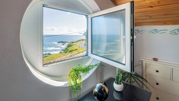 A room with a circular window overlooking a coastal landscape, a small table with decorative items, and a white dresser.