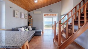 A modern interior with a wooden ceiling, a kitchen area, a grey sofa, and a staircase leading to an outdoor balcony with a view.