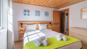 A well-lit bedroom with a wooden headboard, a large bed with a green bedspread, two rolled towels, and decorative tiles on the wall.