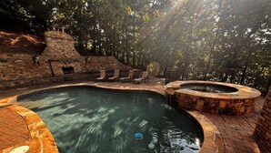 Una piscina con borde de piedra y un jacuzzi, rodeados por un patio de ladrillo y árboles.