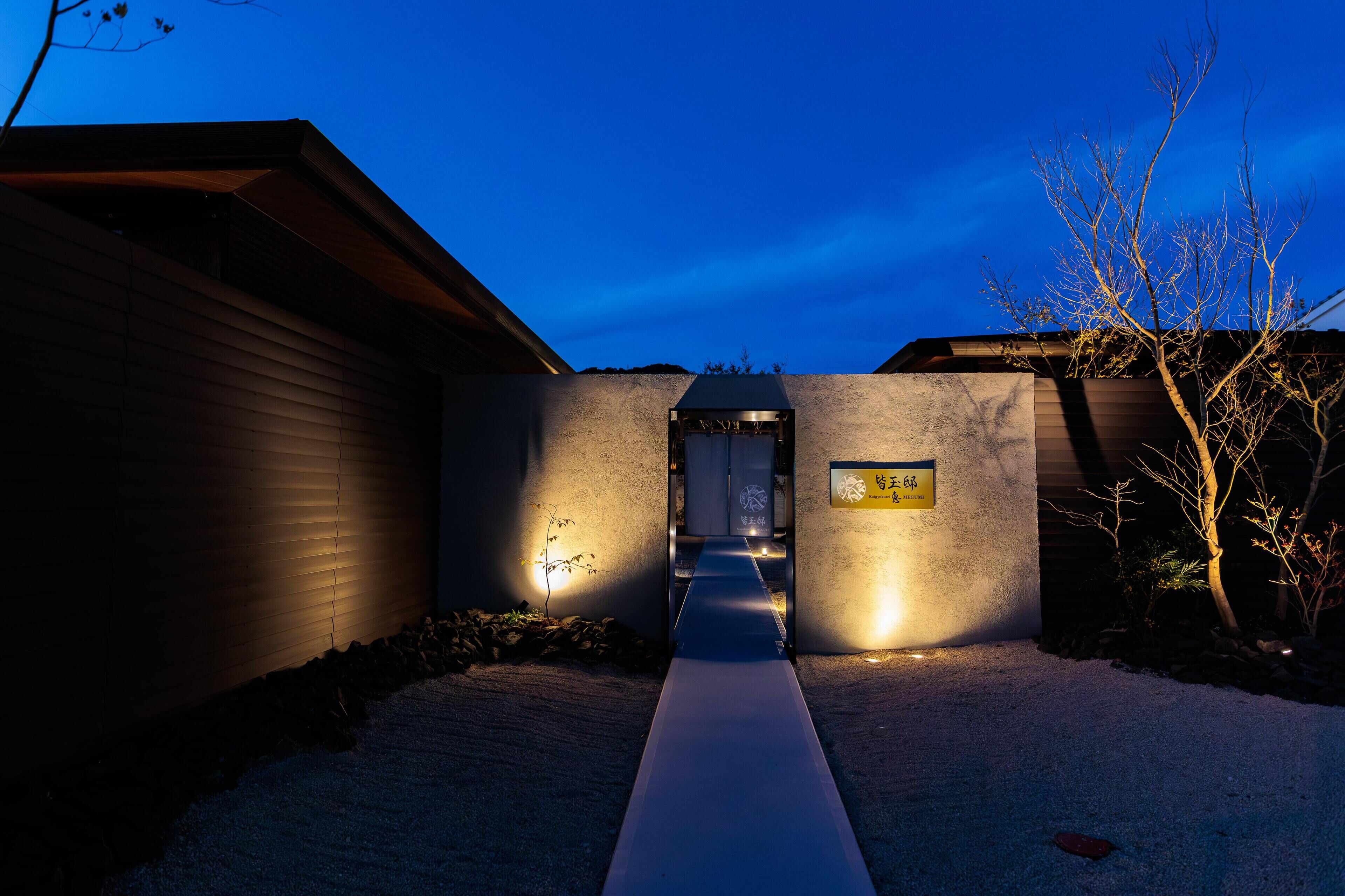 A pathway leading to a building entrance at dusk, with a sign on the wall and a tree to the right.