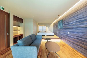 Una habitación de hotel moderna con sofá, una mesita redonda pequeña, una cama con ropa de cama blanca y una pared con textura.