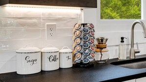 A modern kitchen with white cabinets, a black countertop, and a window with a view of trees. There are three jars labeled 'Sugar', 'Coffee', and 'Tea'.