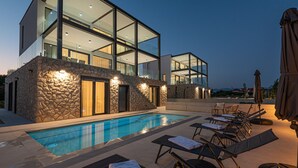 Un bâtiment moderne doté d’une façade en verre, d’une piscine et de chaises longues extérieures.