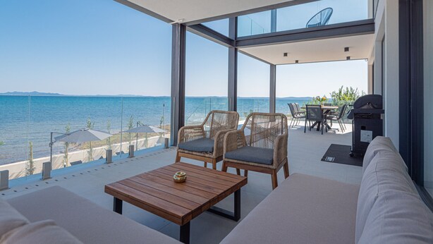 Un espace extérieur moderne, aménagé avec des fauteuils en osier et une table en bois, offrant une vue sur une mer bleue et limpide et sur une côte lointaine.