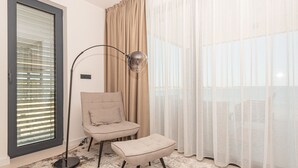 Une pièce comprenant un fauteuil, un pouf, une lampe sur pied et des rideaux légers.