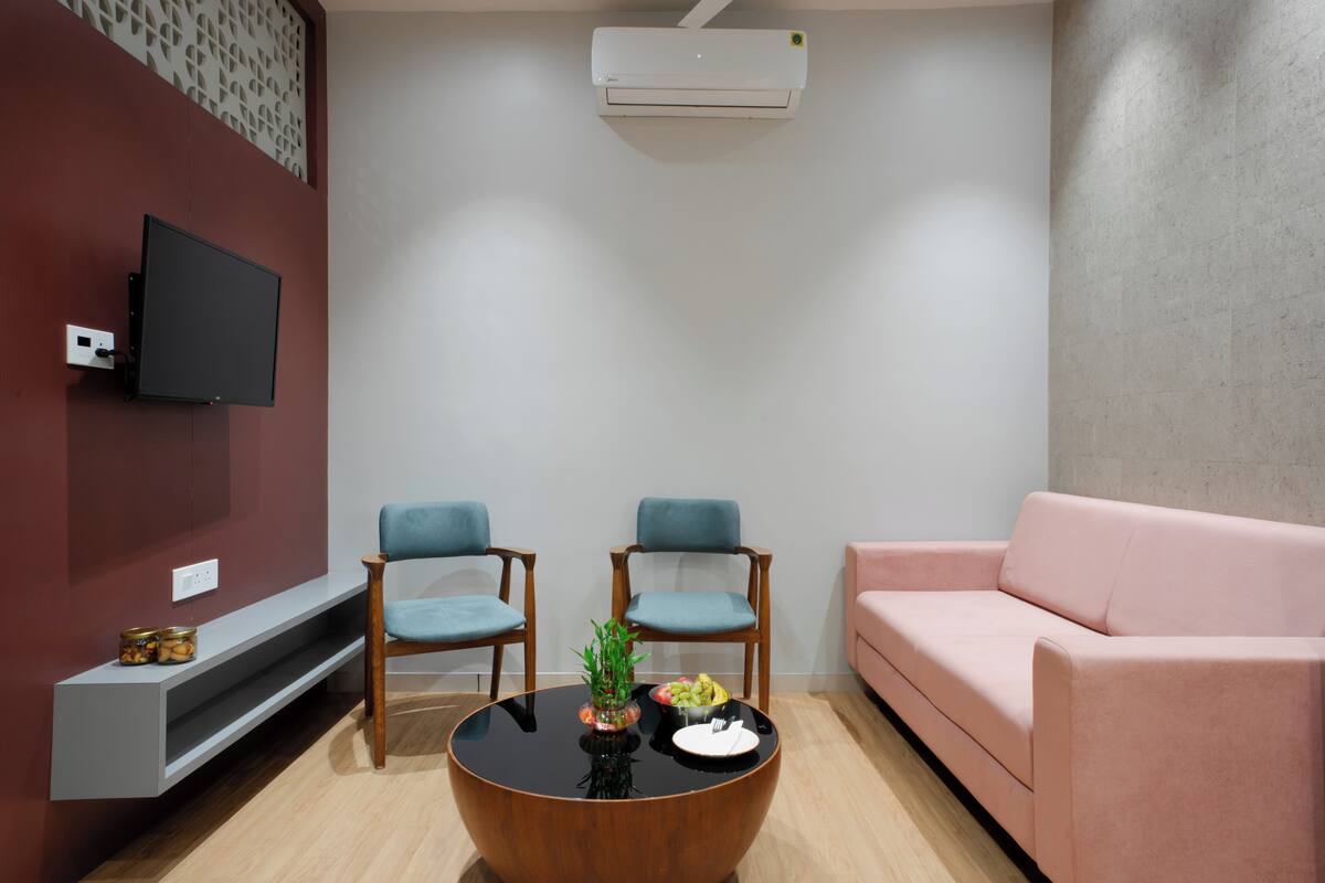Een moderne woonkamer met een flatscreen televisie, twee blauwe stoelen, een ronde houten salontafel met fruit, een roze bank en een vastgehangen airconditioner.