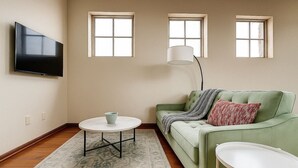 Un salon avec un canapé vert, une table basse blanche, un téléviseur à écran plat et trois fenêtres.