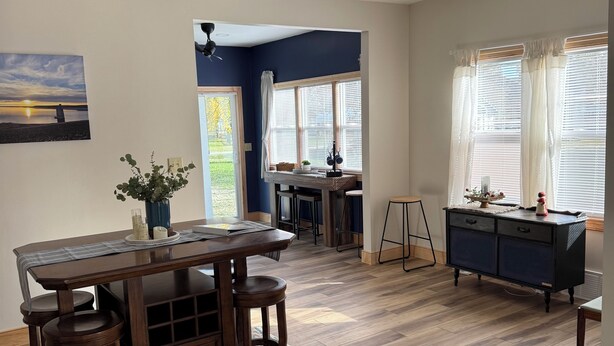 Un comedor amplio con una mesa y sillas de madera, un ventilador de techo y vistas al exterior a través de amplias ventanas.