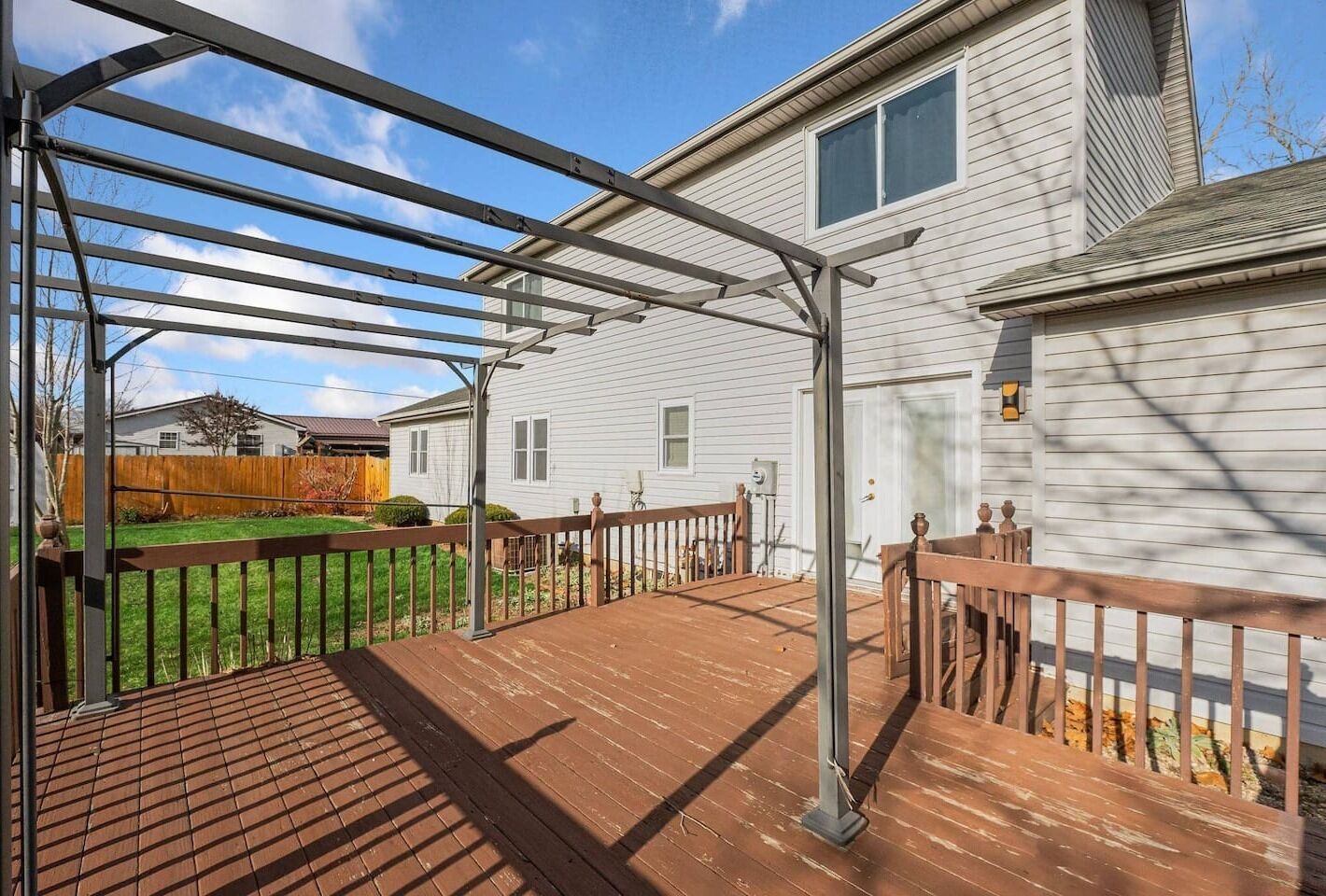 Une terrasse extérieure couverte, dotée d’une structure en métal formant une pergola, d’un revêtement de sol en bois et, au fond, d’une maison blanche.