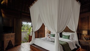 Una cama con dosel y cuatro columnas con sábanas blancas y almohadas verdes, una mesita de noche de madera con una lámpara, y vistas a zonas verdes en el exterior.