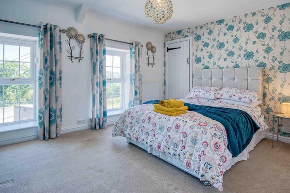 A bedroom with a floral-patterned bedspread, a tufted headboard, and a view of the outdoors through a window with curtains.