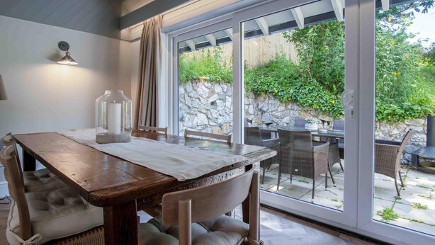 A dining area with a wooden table and chairs, a candle, and a view of the garden through glass doors.
