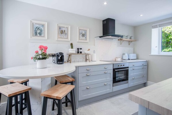 A modern kitchen with a round island, built-in oven, and grey cabinetry. There are three framed pictures on the wall, a potted plant, and a coffee machine on the island.