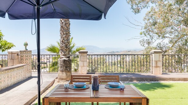 Un espace de restauration en plein air avec une table dressée pour deux personnes, un grand parasol et une vue sur les montagnes.