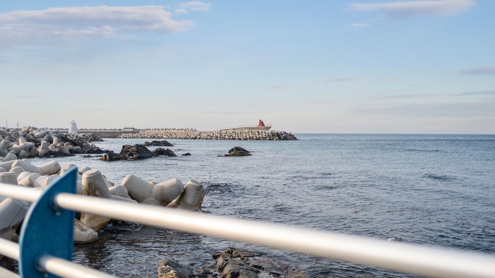 Paesaggio costiero con un faro, frangiflutti rocciosi e mare calmo.