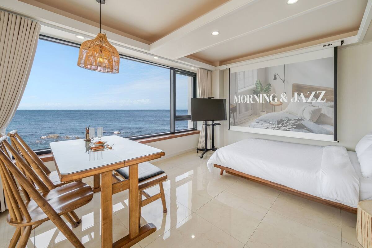 Hotelkamer met een groot raam met uitzicht op zee, een bed, een eettafel met stoelen, een televisie en een kunstwerk aan de muur getiteld 'MORNING & JAZZ'.