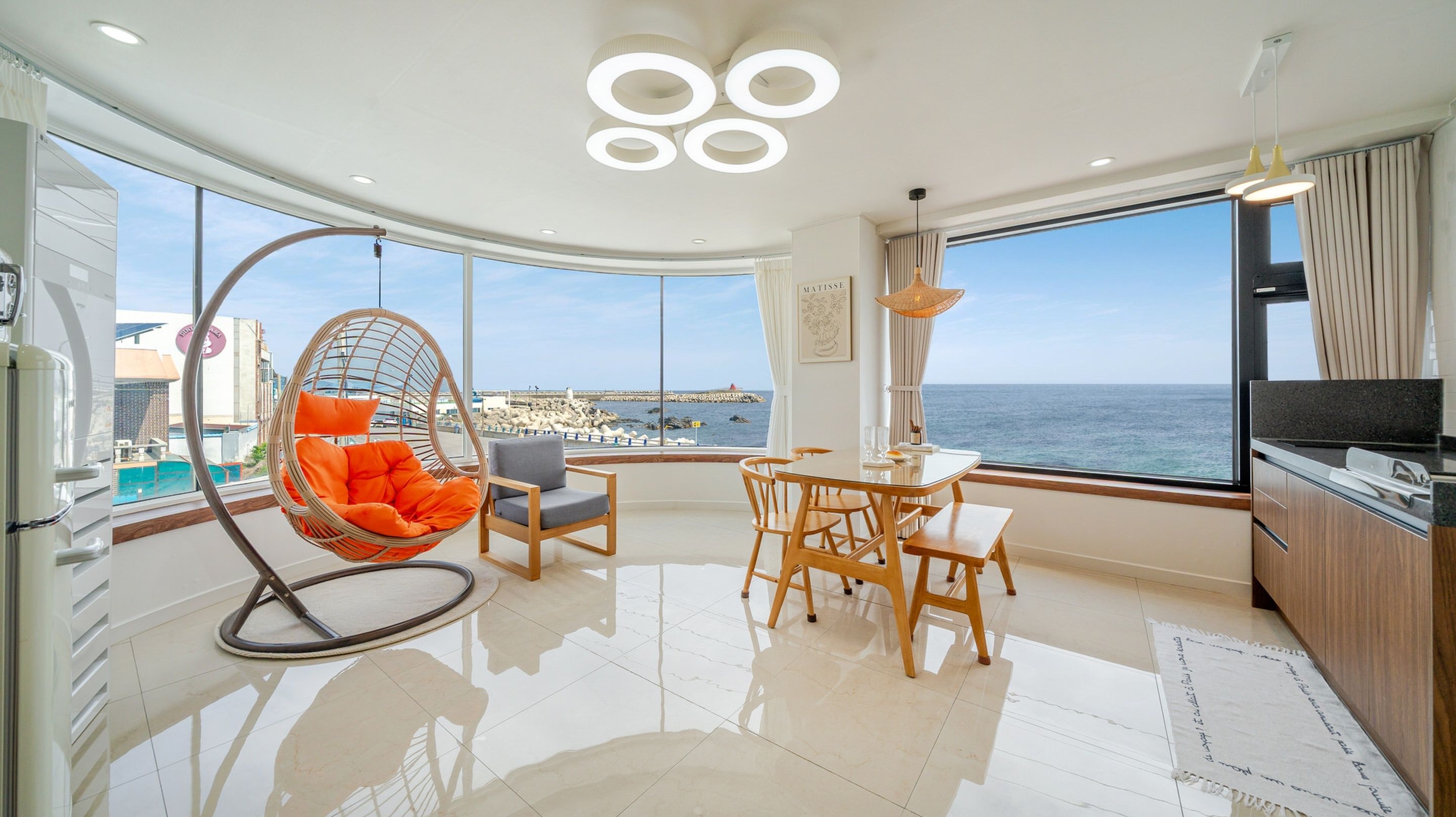 Una sala de estar moderna con un ventanal grande con vista al mar, un comedor con muebles de madera y un sillón colgante con cojines naranjas.