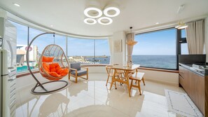 Una sala de estar moderna con un ventanal grande con vista al mar, un comedor con muebles de madera y un sillón colgante con cojines naranjas.