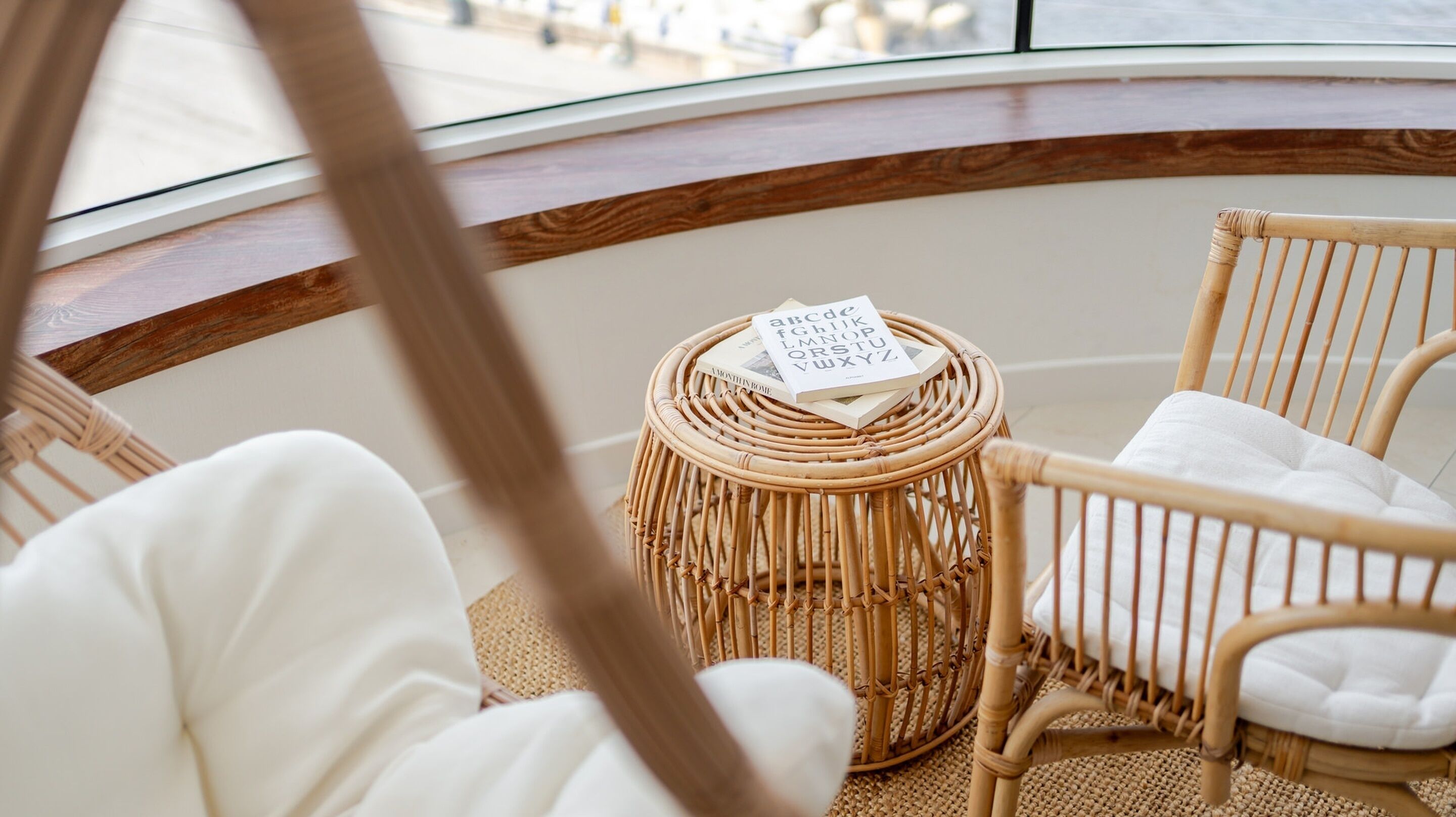 Una silla de mimbre con cojín, una pequeña mesa redonda de mimbre con un libro, y vista al mar a través de una ventana.